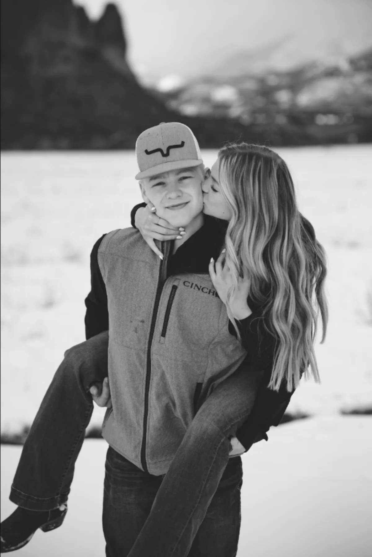 Black and white photo of an engaged couple in a snowy Colorado field, she kisses his cheek while he carries her