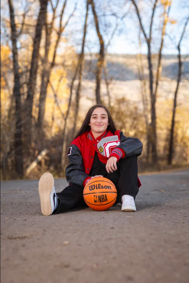 Senior athlete in a red varsity jacket seated with a basketball on an autumn road