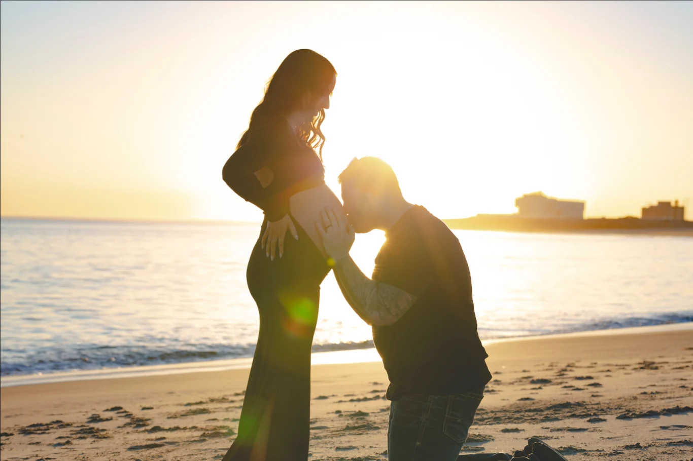Expecting couple silhouetted at sunset on the beach as he kisses her belly