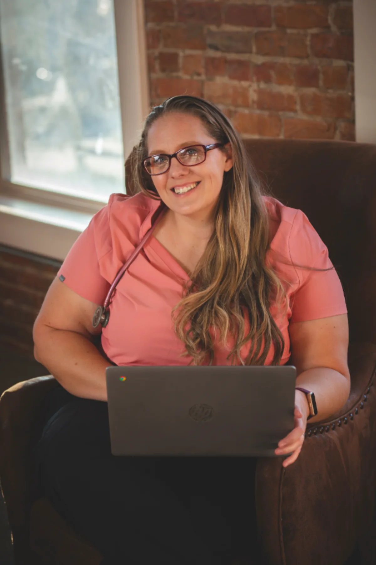 Healthcare provider in pink scrubs seated with a laptop in a brick office