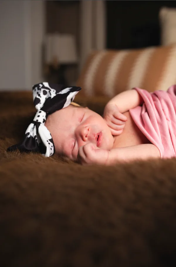 Newborn girl in a pink wrap and patterned bow sleeping on a soft brown blanket at home