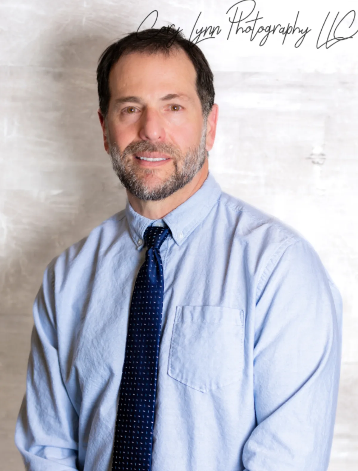 Man in a light blue shirt and navy tie photographed against a textured white wall