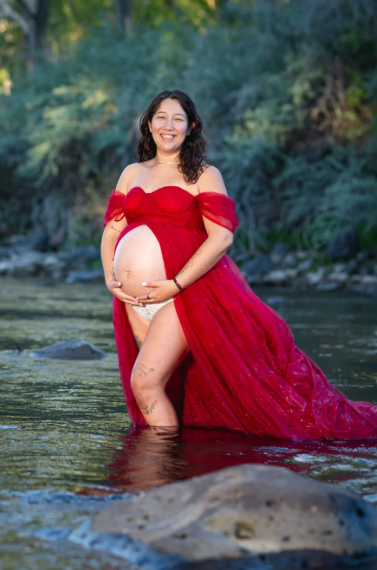 Expecting mother in a flowing red gown standing in a shallow Colorado river