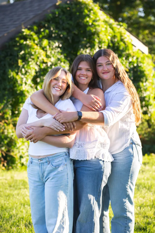 Three sisters laughing while hugging in a green sunlit backyard