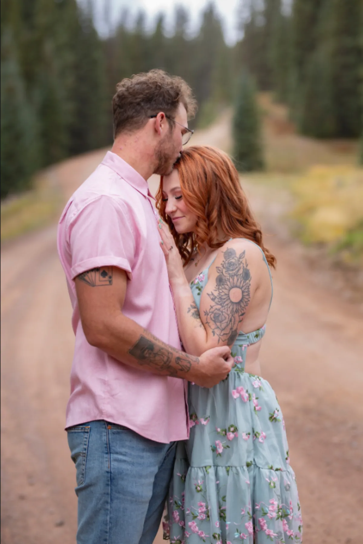 Engaged couple embracing on a pine lined dirt road as he kisses her forehead