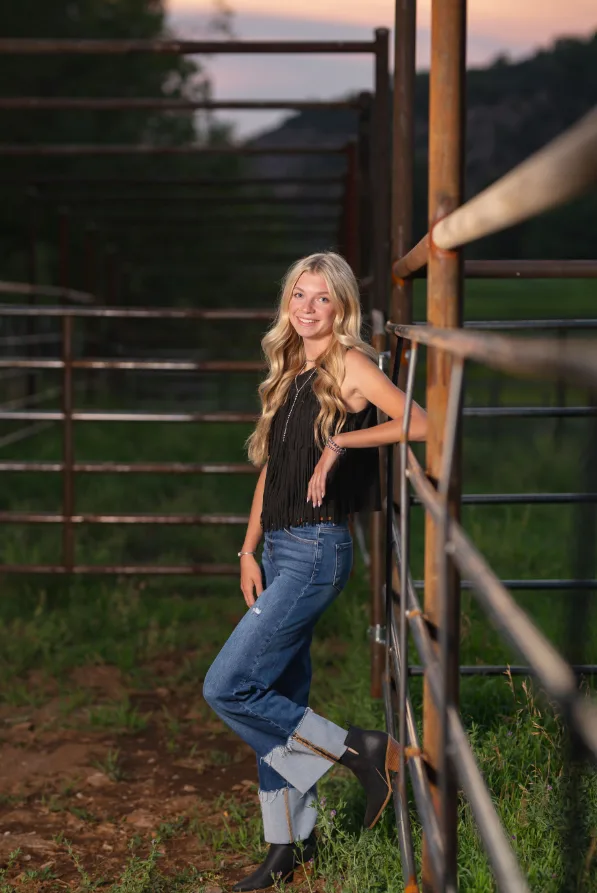 High school senior girl leaning against a ranch corral gate at dusk in Colorado