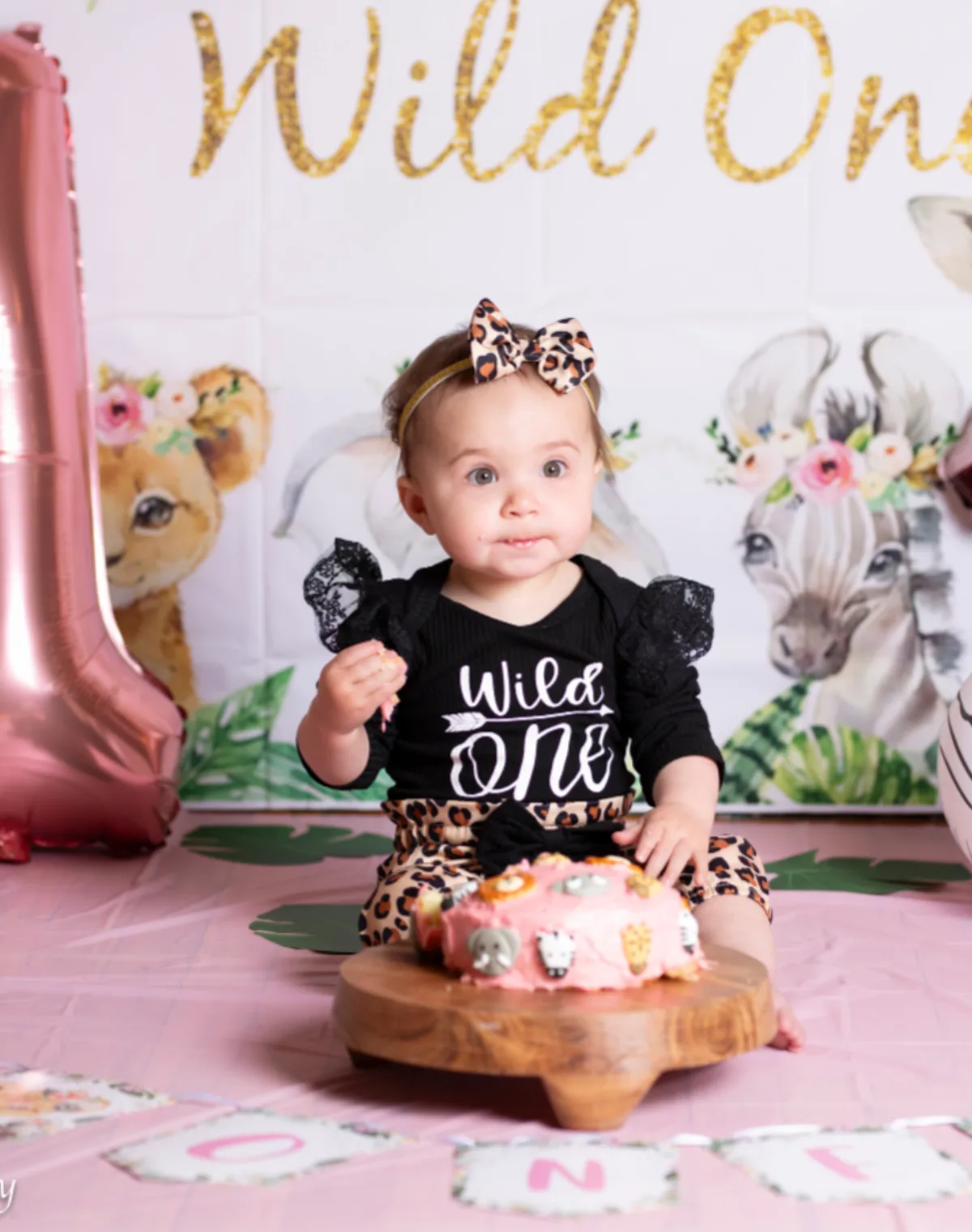 One year old girl in a Wild One outfit sitting beside her pink safari themed smash cake