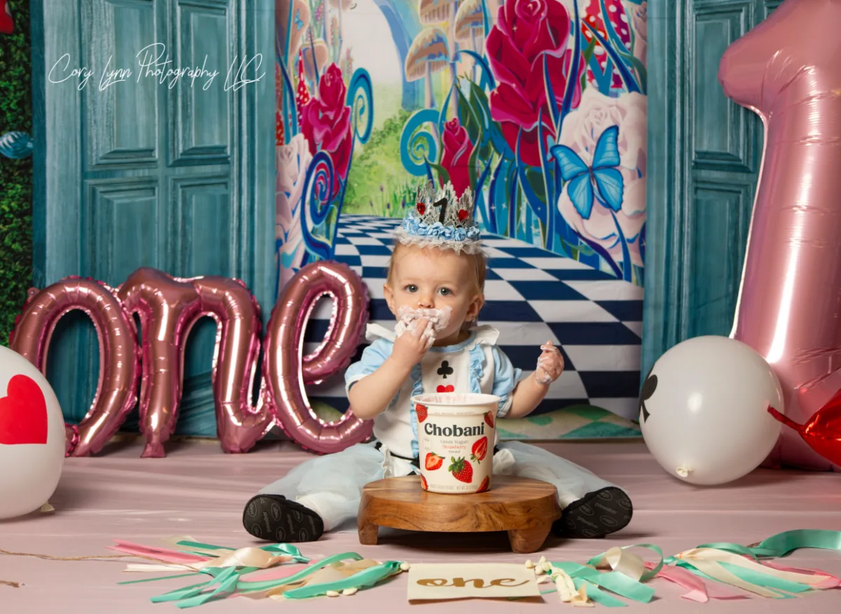 One year old in an Alice in Wonderland outfit covered in frosting in front of a painted garden backdrop