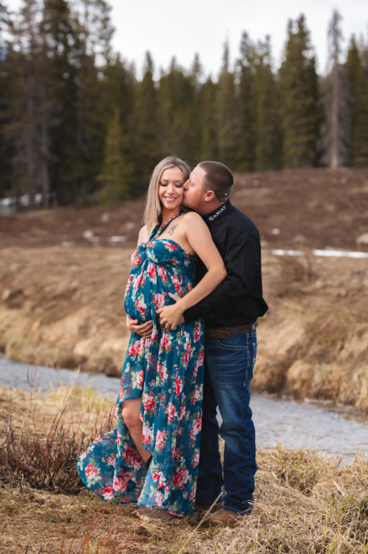 Expecting couple embracing beside a mountain creek with pines behind them