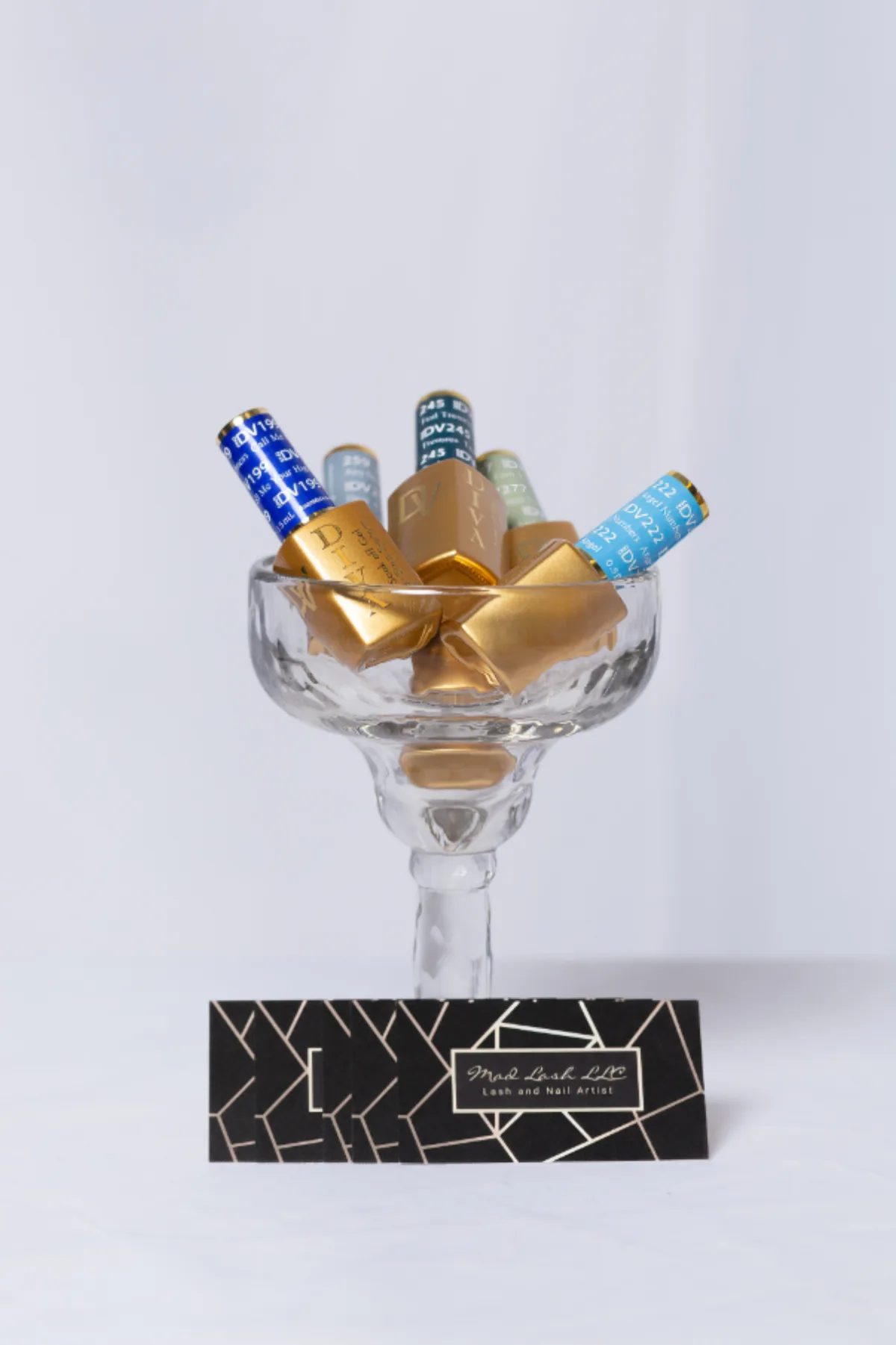 Nail polish bottles arranged in a glass coupe beside a lash and nail artist business card