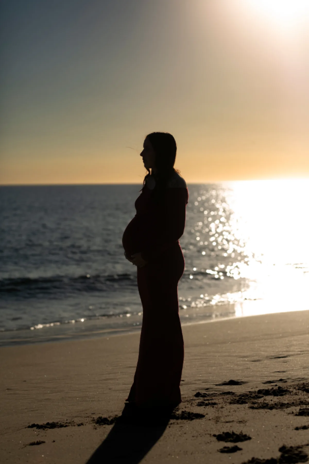 Silhouette of an expecting mother in a long gown standing at the shoreline at sunset