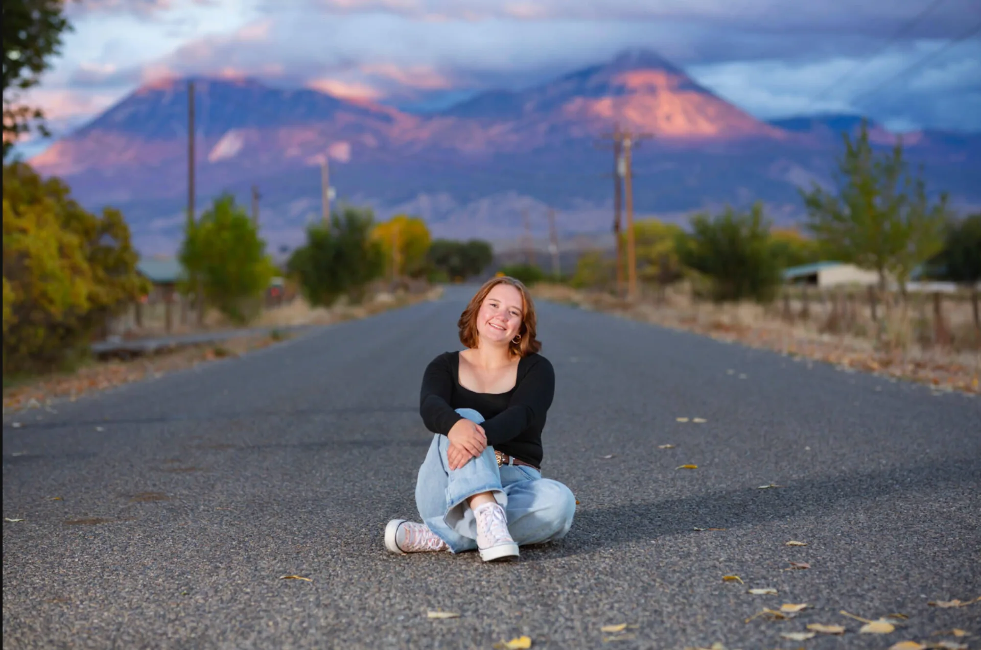 Senior portrait of a girl seated on a rural Colorado road with a mountain range glowing at golden hour