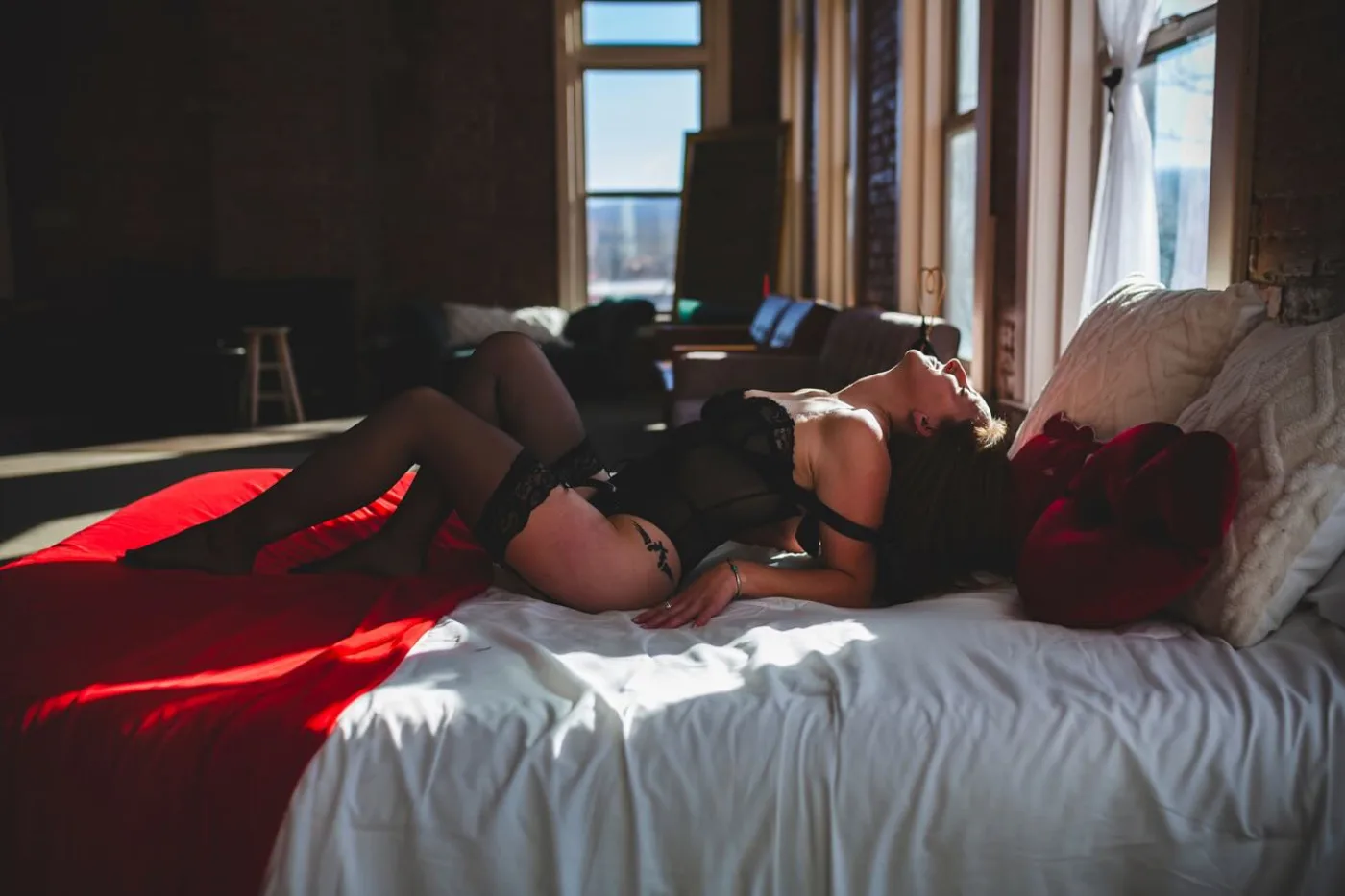 Woman in black lace lingerie reclining on white bedding in a loft flooded with late afternoon sunlight
