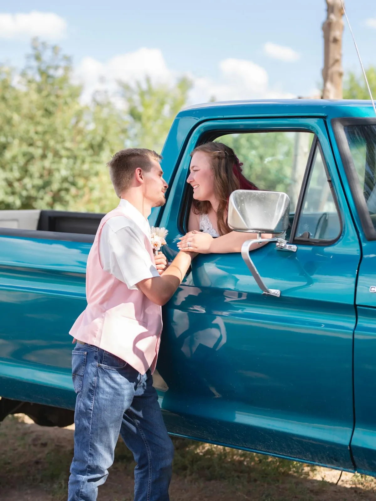 Groom holding his bride's hand as she leans out the window of a vintage teal pickup truck on their wedding day